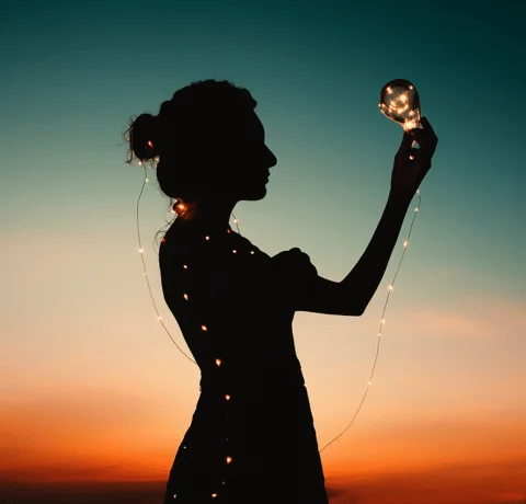 WTribe_–_Voice_&_Vision Silhouette of an Indian woman holding a glowing bulb against a sunset sky, wrapped in string lights.